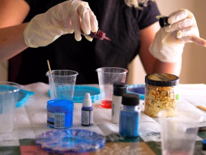 Hands wearing gloves mixing colored epoxy resin with gold flakes and pigments for handmade polymer clay and resin earrings